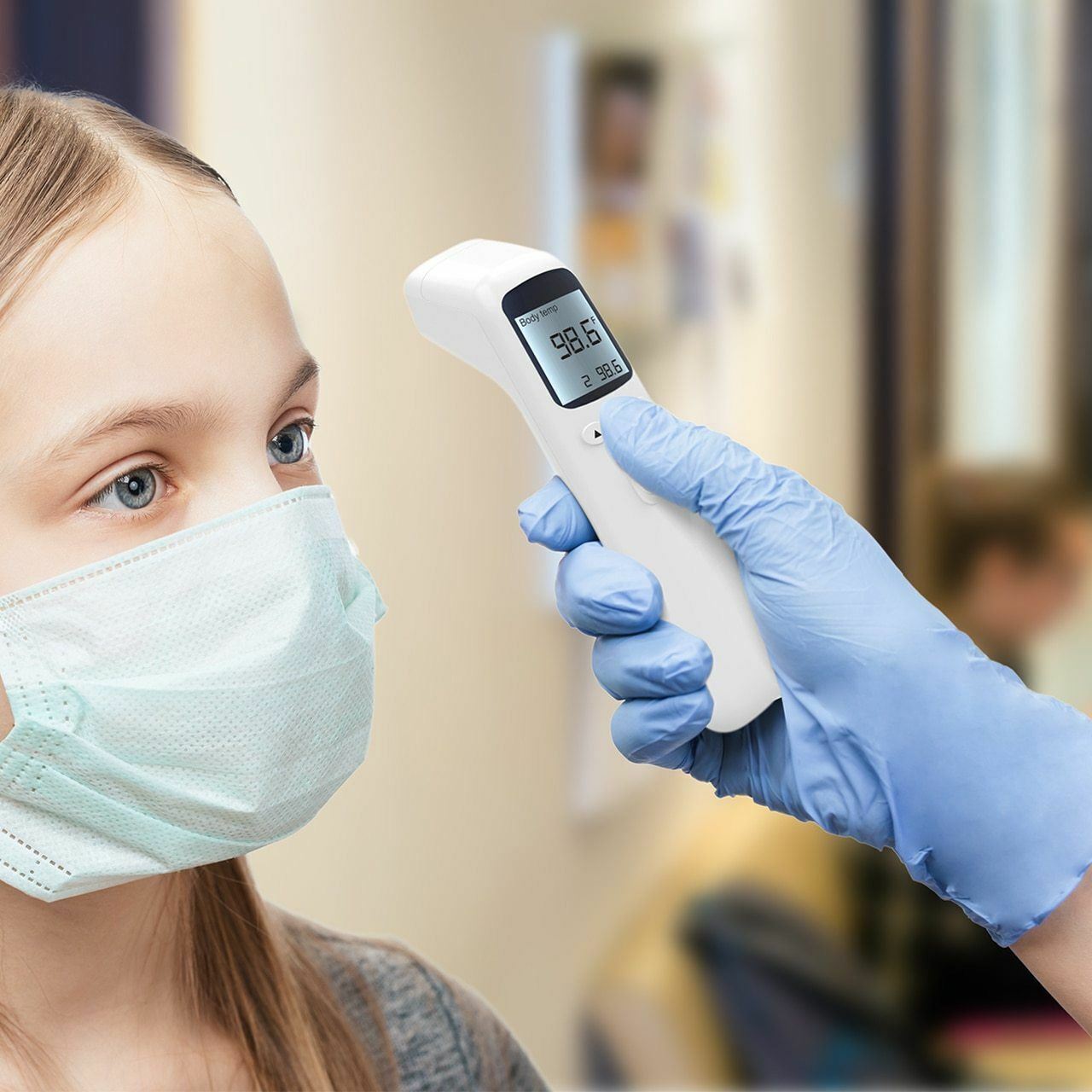 A young girl wearing a surgical mask has her temperature checked by a gloved hand holding a non-contact MediWares Thermometer - Pro Series, which reads 98.6°.